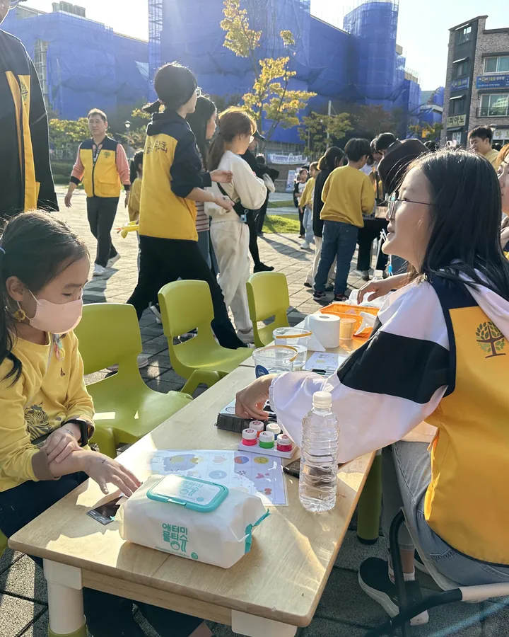축제 천막 부스 테이블에서 노란 조끼를 입은 봉사자와 함께 체험을 하는 아이
