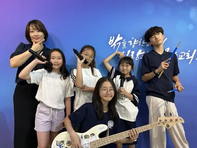 파란 배경막 앞에서 베이스 기타·드럼 스틱을 든 아이들과 선생님