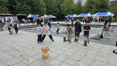 천막 부스가 늘어선 공원 광장에서 아이들이 투호 놀이를 하는 축제 풍경
